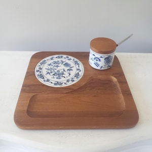 May include: A square wooden serving tray featuring a circular white ceramic inlay with a blue floral pattern. A matching cylindrical ceramic container with a wooden lid and a small metal spoon sits on the tray, alongside a carved rectangular section.