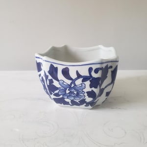 May include: A small, white ceramic bowl with a blue floral design. The bowl has a unique, scalloped edge and a square shape. The blue floral pattern is detailed and covers the exterior of the bowl, creating a classic, decorative piece.
