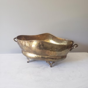 May include: A hammered brass basin with an oval shape, featuring a wavy rim, two side handles, and four decorative feet. The metal surface has a polished, reflective finish with visible indentations from the hammering process throughout.