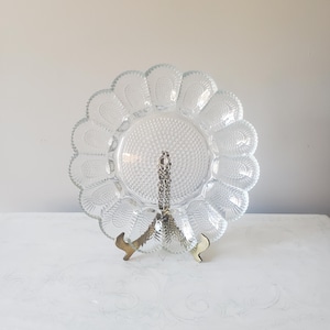 May include: A clear glass serving dish with a scalloped edge and a central section with a textured pattern. The dish is supported by a gold-colored metal stand. The dish is designed to hold appetizers or desserts.
