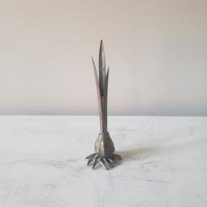May include: A metallic sculpture shaped like a sprouting bulb with a rounded base, radiating root-like structures at the bottom, and three tall, pointed, leaf-like blades extending vertically from the center, featuring a weathered, tarnished finish.