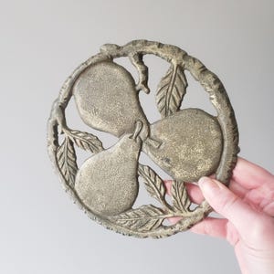 May include: A round, antique-style trivet with a pear design. The trivet is made of a metallic material with a weathered, bronze finish. Three pears are arranged in the center, surrounded by leaves and branches. The trivet is held by a hand.
