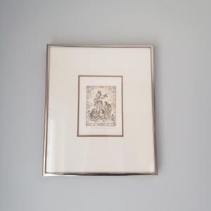 May include: Framed artwork featuring a sepia-toned illustration. The artwork depicts a central figure with several smaller figures below. The frame is silver-colored, and the artwork is set against a white mat.