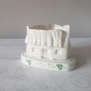 May include: A cream-colored ceramic cottage-shaped planter or trinket holder featuring a textured thatched roof, embossed windows, and a door. The base is decorated with a raised lattice pattern and hand-painted green shamrock accents.