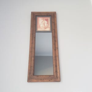 May include: A tall, rectangular mirror with a distressed, brown wooden frame. The top section features a framed sepia-toned print of two figures. The mirror's reflective surface fills the lower portion, offering a clear reflection. The mirror is mounted on a light gray wall.