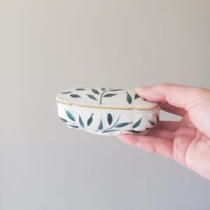 May include: An oval ceramic trinket box with a white base and a hand-painted dark green leaf pattern. The lid features a scalloped edge with a thin gold trim, and the body of the box has a matching scalloped design along the bottom rim.