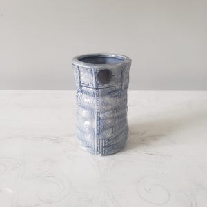 May include: A cylindrical ceramic vase with a light blue glaze and a textured surface that mimics the appearance of denim fabric, complete with molded details resembling stitched seams, pockets, and a dark circular button on the upper front.