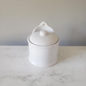 May include: A white ceramic wall-mounted salt cellar featuring a cylindrical base, a removable lid with a small knob handle, and a back panel with a single hole for hanging. The container has a smooth, glossy finish and a simple, functional design.