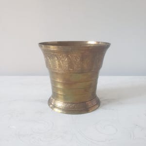 May include: A brass mortar with a wide, flared rim and a tapered body. The upper section features a band of etched leaf patterns, while the lower section is decorated with horizontal lines and a base band of repeating leaf motifs.