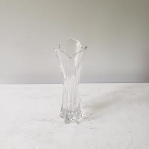 May include: A clear glass vase with a flared, tulip-shaped top and a textured, ribbed body. The vase has a star-shaped base and stands on a white surface. The vase is designed to hold flowers and other decorative items.