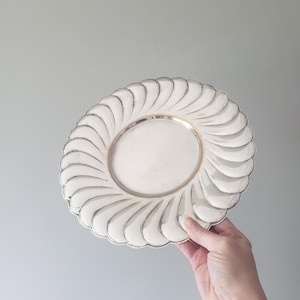 May include: A round silver-toned plate featuring a smooth, flat center surrounded by a wide, decorative border with a repeating, embossed swirl pattern that radiates outward toward the scalloped, curved edge of the metallic serving dish.