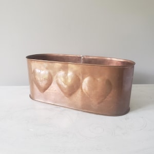 May include: An oval copper planter featuring three embossed heart shapes along the front side. The metallic container has a smooth, polished finish and a wide, open top, showcasing a warm, reddish-brown tone throughout the entire exterior surface.