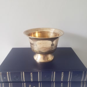 May include: A small, gold-colored metal bowl with a rounded rim and a pedestal base. The bowl has horizontal bands around the middle and sits on top of two dark blue books.