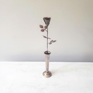 May include: A silver metal vase holds a metal rose with detailed leaves and petals. The rose is a dark, metallic color, and the vase has a flared base and a slender neck. The background is a light gray.