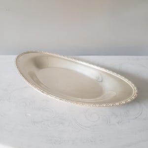 May include: An oval-shaped silver-toned serving tray with a smooth, reflective interior surface and a decorative, textured beaded border along the outer rim. The tray features a shallow basin and a polished metallic finish throughout its structure.