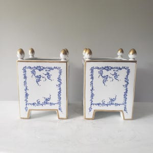 May include: A pair of square white ceramic planters featuring blue floral patterns framed by thin blue lines. Each planter has four short legs and four decorative finials on the top corners, all accented with gold trim along the edges and details.