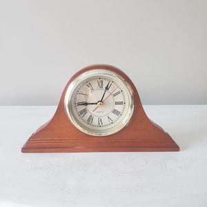 May include: A wooden mantel clock with a gold-colored face and black Roman numerals. The clock has a brown wooden base and a curved top. The clock hands are black and red. The clock face reads "QUARTZ".