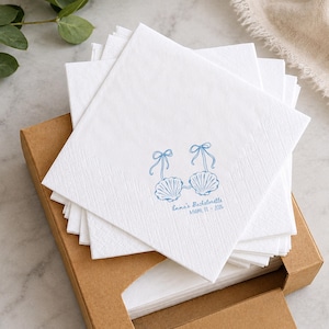 May include: A stack of square white paper napkins featuring a blue graphic of two seashell bikini tops with ribbons. The text printed in blue below the shells reads Emma's Bachelorette MIAMI, FL - 2026. The napkins are inside an open brown cardboard box.