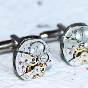 May include: A pair of silver cufflinks with intricate watch mechanisms. The cufflinks feature exposed gears and a silver finish.