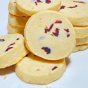 May include: A stack of round, light-colored cookies with visible pieces of dried cranberries. The cookies are arranged on a white surface, with some cookies stacked and others scattered around. The cookies have a slightly rough texture.