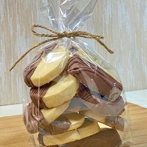 May include: A clear cellophane bag tied with twine, filled with shortbread cookies dipped in chocolate. The cookies are a light tan color, with a semi-circle of dark brown chocolate on top. The bag is sitting on a wooden surface.