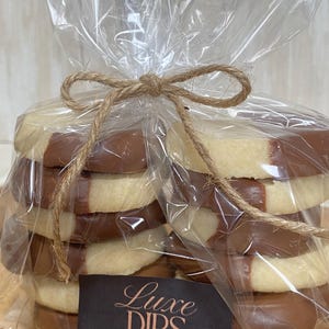 May include: A stack of shortbread biscuits, half-dipped in chocolate, are packaged in clear cellophane and tied with twine. The biscuits are from Luxe Dips Sweet Shop.