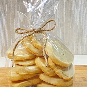 May include: A clear cellophane bag tied with twine contains a stack of round, golden-brown cookies. The cookies have a slightly uneven surface and are stacked neatly. The twine is tied in a bow at the top.