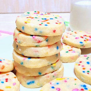 May include: A stack of round, light-colored cookies with colorful sprinkles. Several cookies are scattered around the stack. A glass of milk is in the background. The cookies appear to be shortbread or sugar cookies.