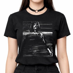 Può includere: T-shirt nero con grafica in bianco e nero di una persona seduta. La modella indossa un choker nero e una gonna a pieghe nera.