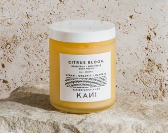 May include: A jar of Citrus Bloom body polish with a white lid and a label. The label includes the product name, ingredients, and brand name. The jar is sitting on a light-colored rock against a textured background.
