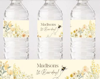 Op de afbeelding: Drie doorzichtige waterflessen met crèmekleurige etiketten. De etiketten tonen bloemmotieven in geel en wit, en de tekst "Madisons 1st Bee-day!" met een bijengrafiek. De flessen hebben witte doppen.