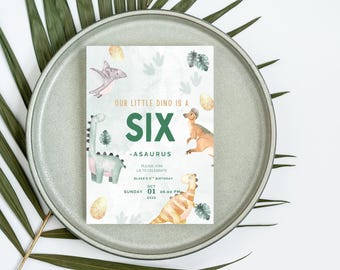 May include: A birthday invitation with a dinosaur theme. The invitation features watercolor illustrations of dinosaurs, eggs, and foliage. The text reads "OUR LITTLE DINO IS A SIX -ASAURUS" with the date and time of the party. The invitation is on a gray plate.