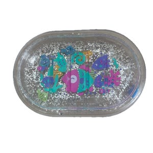 May include: A clear, oval-shaped soap dish filled with silver glitter and colorful sea creatures. The sea creatures include fish in shades of pink, blue, green, and purple. The dish is made of clear plastic.