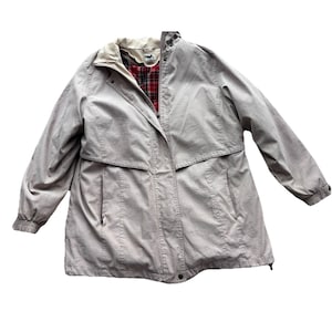 May include: A light tan, long coat with a beige collar and a red and black plaid lining. The coat has a zipper closure, two front pockets, and elastic cuffs. The coat is a neutral color and suitable for various outfits.