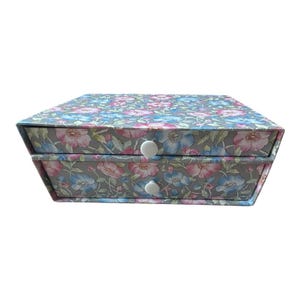 May include: A rectangular storage box with two stacked drawers, each featuring a small white circular knob. The exterior is covered in a gray floral pattern with pink and blue flowers and green leaves throughout the entire surface.