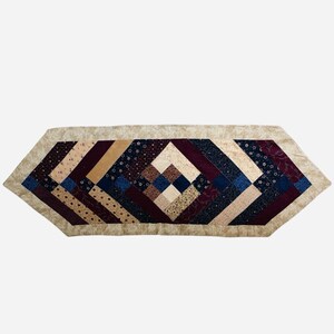 May include: A quilted table runner with a geometric pattern. The runner features a beige border and a central design of diamond shapes in burgundy, navy, tan, and cream fabrics. The patchwork design adds a rustic touch.