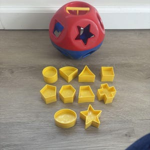 May include: A red, blue, and yellow shape sorter toy with various geometric shapes. The toy includes a red ball with different shaped holes and ten yellow shapes, including circles, triangles, and stars. The shapes are numbered.