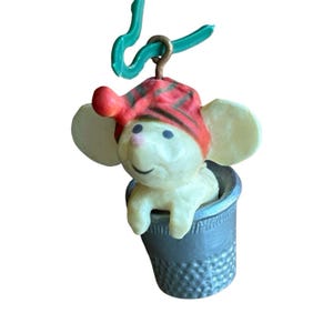 May include: A small cream-colored mouse figurine wearing a red and black plaid hat, positioned inside a metallic gray thimble. The mouse has large ears, a pink nose, and a green plastic hook attached to the top for hanging purposes.