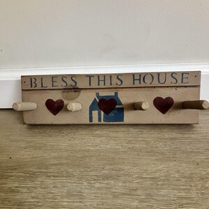 Könnte beinhalten: Eine rechteckige Wandhakenleiste aus Holz mit hellbrauner Oberfläche. Der Text BLESS THIS HOUSE ist in Dunkelblau aufgedruckt. Das Design zeigt drei dunkelrot bemalte Herzen und eine blaue Haussilhouette sowie vier Holzhaken zum Aufhängen.