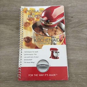 May include: A spiral-bound instruction and recipe booklet for a KitchenAid stand mixer. The cover features a red mixer, a stainless steel bowl with batter, and cupcakes. Text includes Instructions and Recipes for your KitchenAid Stand Mixer.