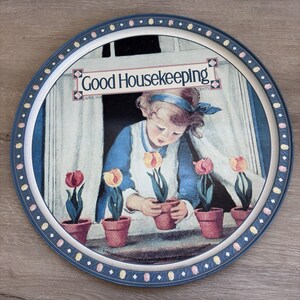 Puede incluir: Bandeja decorativa redonda con una ilustración vintage de una niña cuidando tulipanes en macetas. La bandeja tiene un borde azul con un patrón floral y el texto "Good Housekeeping".