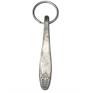 Puede incluir: Un llavero de metal plateado que presenta un mango de cuchara reutilizado como colgante. El mango tiene una forma cónica y alargada con un patrón decorativo en relieve de flores y volutas en el extremo inferior y una anilla metálica redonda.