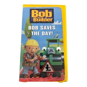Könnte beinhalten: Eine gelbe VHS-Kunststoffhülle mit dem Titel Bob the Builder und dem Text Bob Saves the Day! Das Cover zeigt eine Zeichentrickfigur mit gelbem Helm, eine grüne Straßenwalze, ein Verkehrsschild und vier kleine Igel auf einem roten Untergrund.