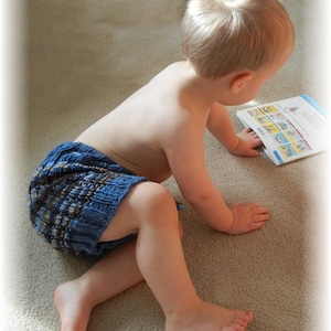 May include: A young child wearing a blue and brown knitted diaper cover sits on a beige carpet and reads a book with colorful illustrations.