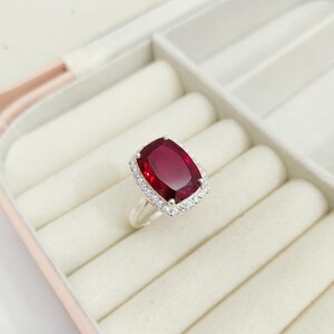 Puede incluir: Un anillo de plata con una gran gema rectangular de color rojo intenso. La gema est&aacute; rodeada por un halo de piedras m&aacute;s peque&ntilde;as y transparentes. El anillo se exhibe en un joyero rosa y blanco.
