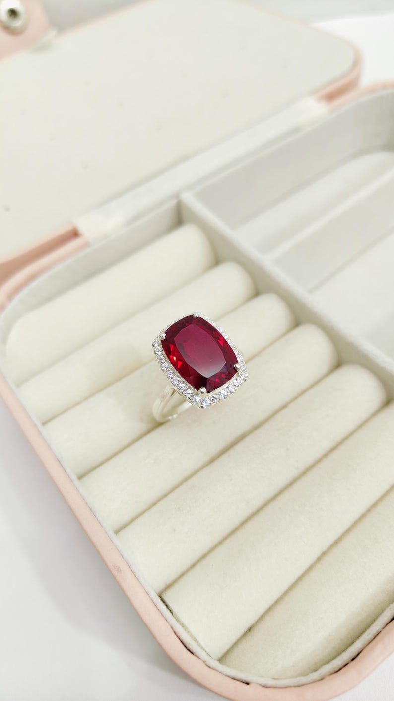 Puede incluir: Un anillo de plata con una gran gema rectangular de color rojo intenso. La gema est&aacute; rodeada por un halo de piedras m&aacute;s peque&ntilde;as y transparentes. El anillo se exhibe en un joyero rosa y blanco.