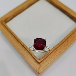 May include: A silver ring featuring a large, square-cut, deep red gemstone. The ring is set on a light-colored background, inside a wooden frame. The gemstone's color is a rich, vibrant shade of red.