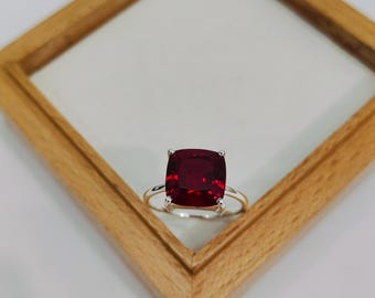 Puede incluir: Un anillo de plata con una gran gema roja oscura de talla cuadrada. El anillo está colocado sobre un fondo claro, dentro de un marco de madera. La gema es de un rojo intenso y vibrante.