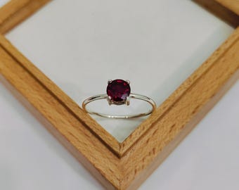 Puede incluir: Un anillo de plata con una gema redonda de color rojo intenso. El anillo está sobre un fondo blanco y enmarcado por un cuadrado de madera.