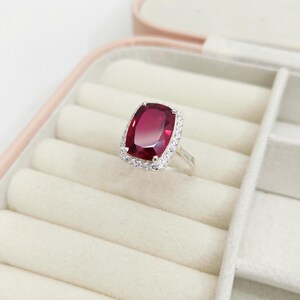 Puede incluir: Un anillo de plata con una gran gema rectangular de color rojo intenso. La gema est&aacute; rodeada por un halo de piedras m&aacute;s peque&ntilde;as y transparentes. El anillo se exhibe en un joyero rosa con interior blanco.